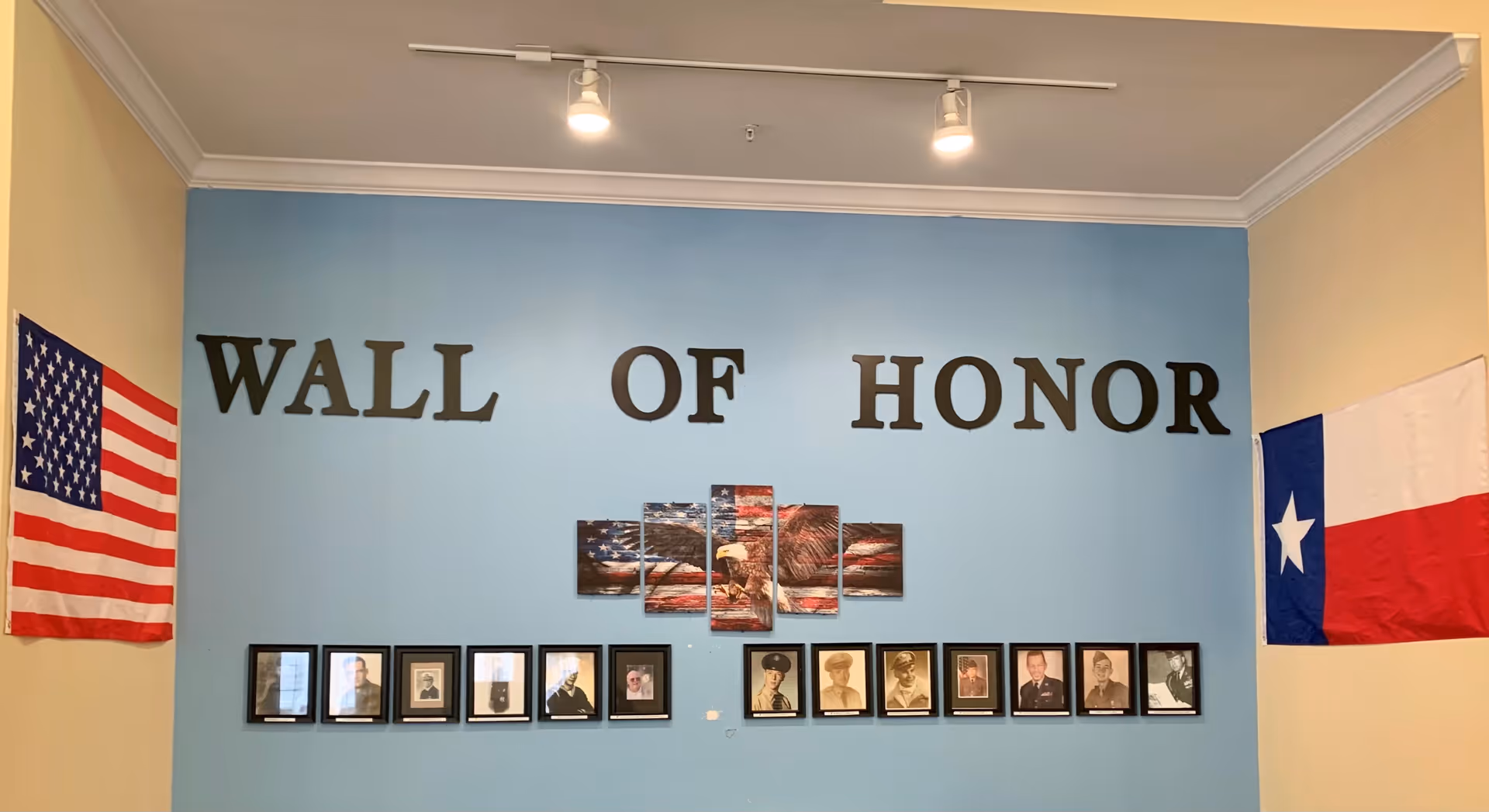A Wall of Honor display with framed photos of veterans beneath a multi-panel artwork of a bald eagle and American flag. The wall is painted light blue with large black letters spelling 'WALL OF HONOR'. An American flag hangs on the left side and a Texas state flag hangs on the right side of the wall.