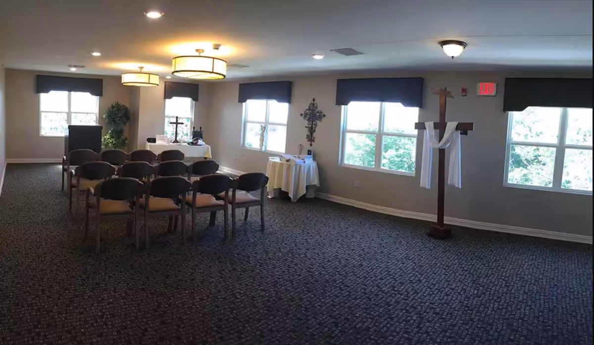 A small chapel or prayer room with rows of chairs facing a table with religious items, including crosses. The room has several windows with black valances, carpeted floor, and ceiling lights. A wooden cross draped with white cloth stands near the windows.