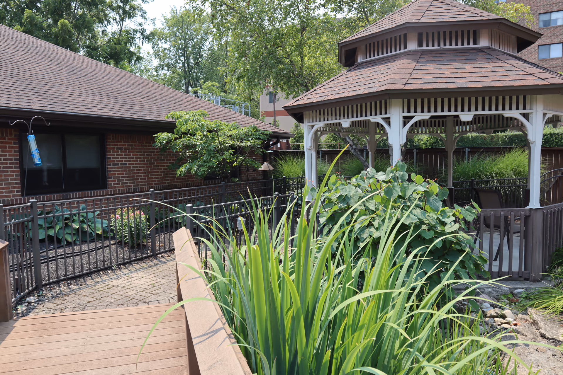 Outdoor garden area at Laurels of Kettering featuring a wooden gazebo with a shingled roof, surrounded by green plants and bushes. There is a brick building with a window and a black metal fence enclosing the garden space. A bird feeder hangs near the window.