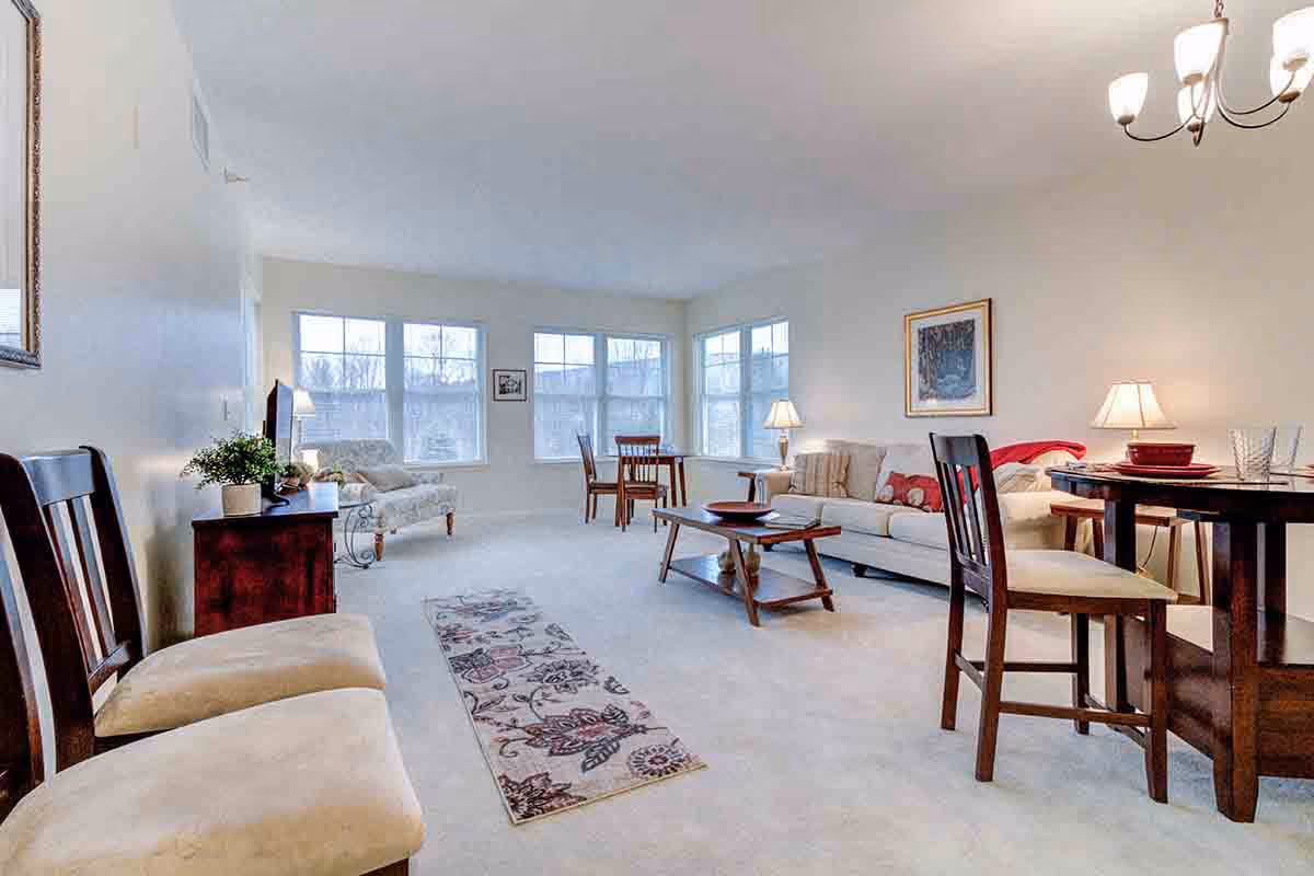 A bright and spacious living room with large windows letting in natural light. The room features a beige carpet, a floral patterned rug, a beige sofa with cushions, a wooden coffee table, a small dining table with chairs, and additional seating including a cushioned bench and an armchair near the windows. There are table lamps and a chandelier providing warm lighting, and a framed picture hanging on the wall.