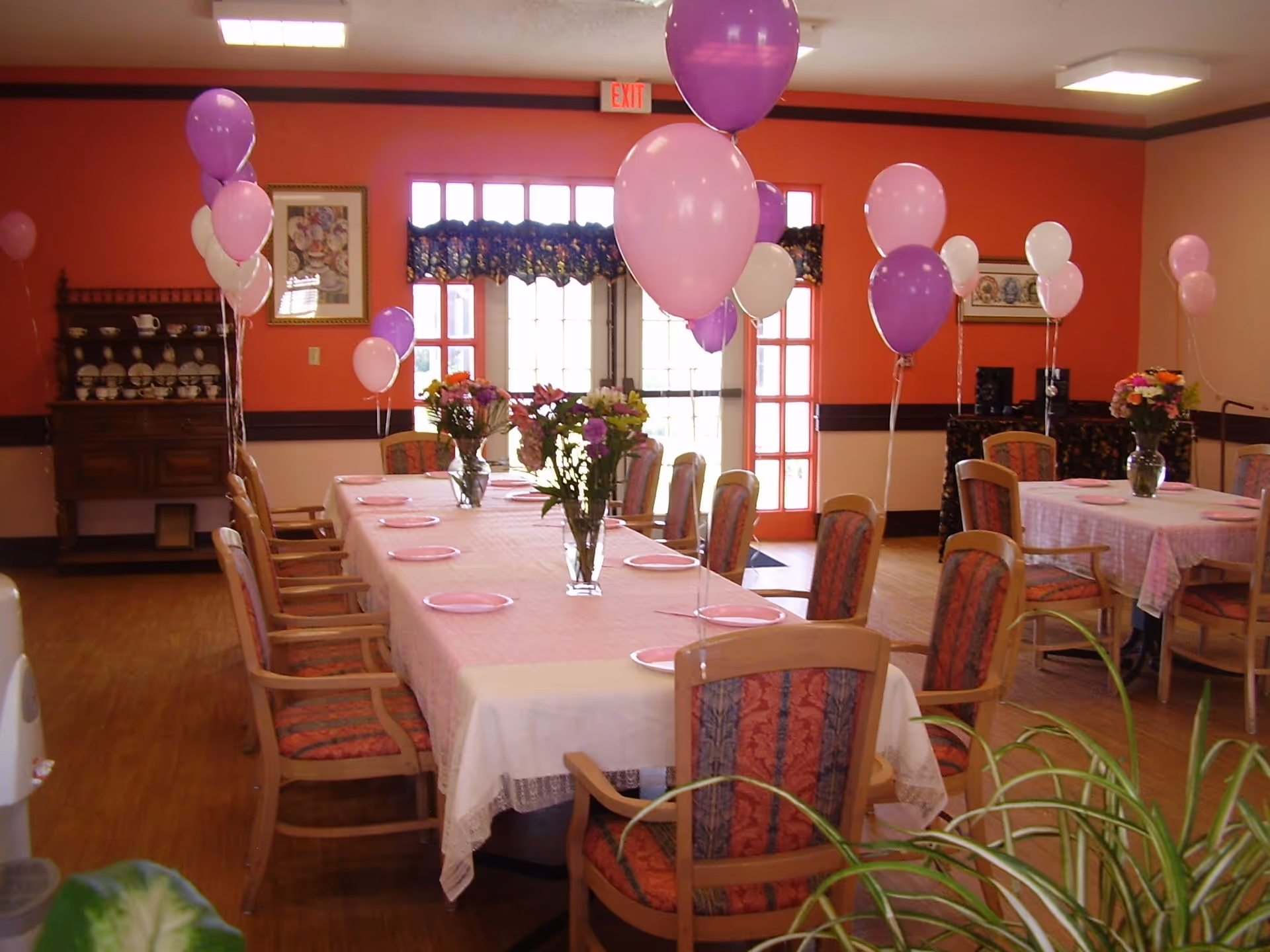 A decorated dining room with tables covered in pink tablecloths, pink plates, and vases of flowers. Purple, pink, and white balloons are tied to the chairs. The room has wooden chairs with patterned cushions, orange walls, and large windows with floral valances letting in natural light.