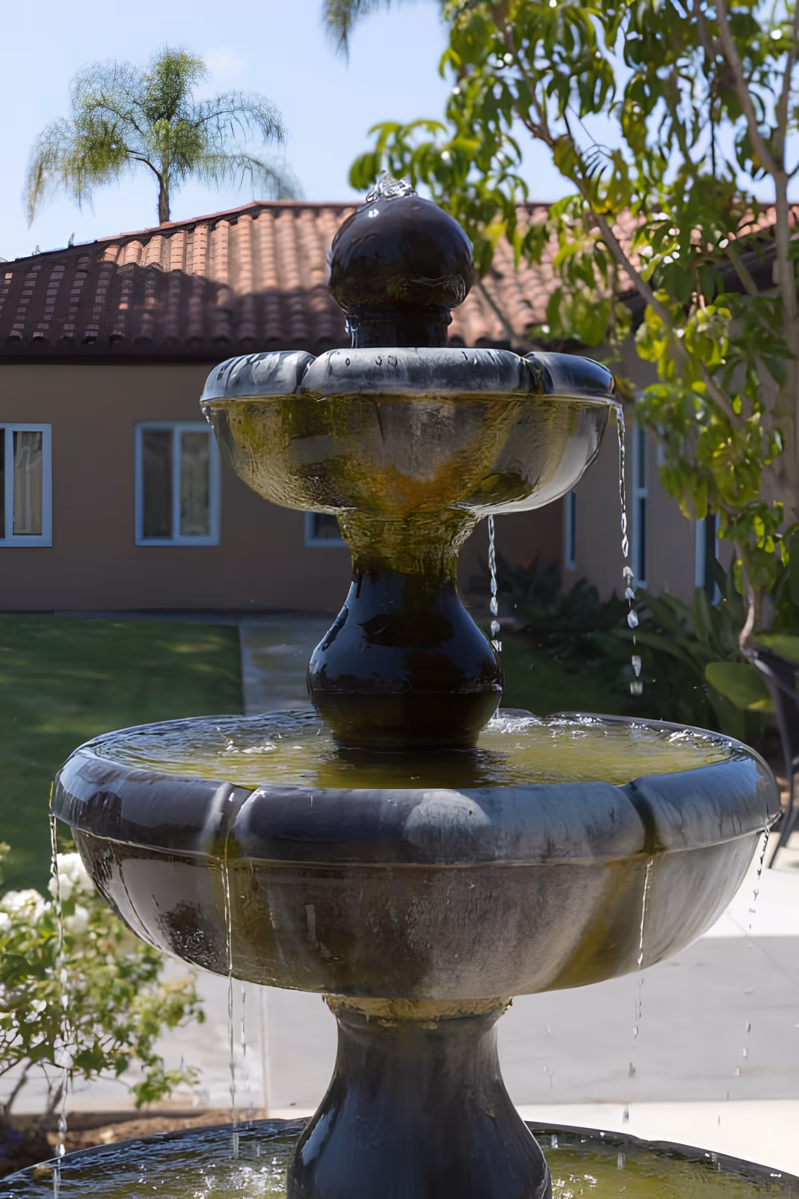 A close-up view of a three-tiered water fountain with water flowing from the top tier to the lower tiers, set outdoors with a building with a tiled roof and windows in the background, along with green trees and plants.