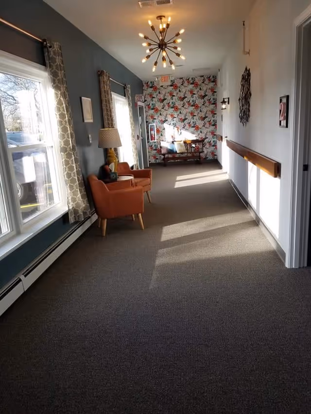 A well-lit hallway in a senior living facility with large windows on the left side covered by patterned curtains. There are two orange armchairs with a lamp and a small table between them along the left wall. At the end of the hallway, there is a bench with colorful cushions against a floral wallpapered wall. The right wall is white with a handrail and some wall decorations. A modern chandelier hangs from the ceiling.