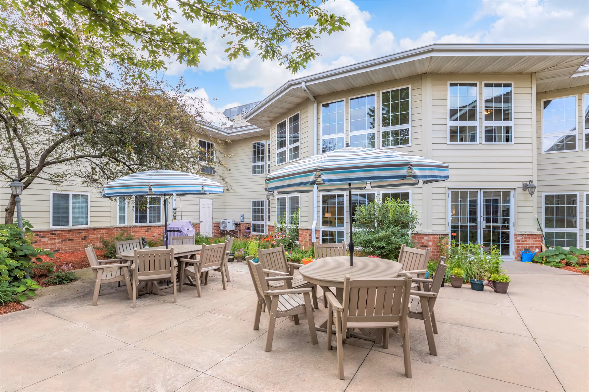Outdoor patio area at a senior living facility with two round tables, each surrounded by six beige chairs and shaded by blue and white striped umbrellas. The patio is surrounded by a beige building with large windows and some greenery including potted plants and trees.