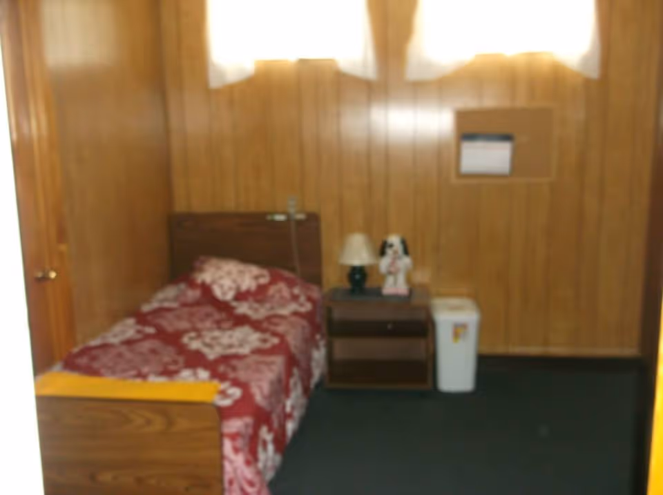 A small bedroom with wood-paneled walls, a single bed with a red and white floral bedspread, a wooden nightstand with a lamp and a decorative dog figurine, a white trash bin, and a window with white curtains.