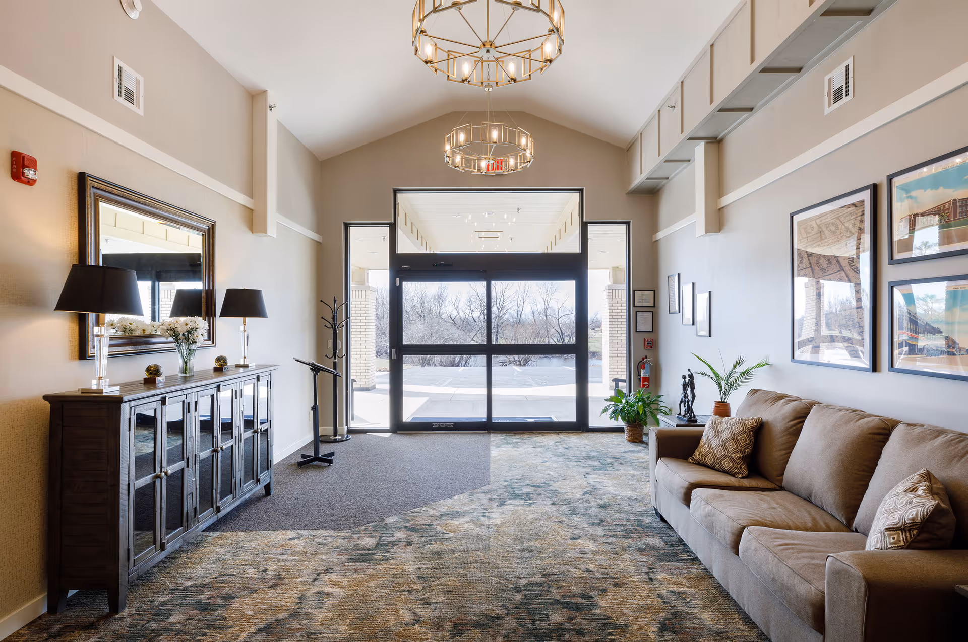 Interior view of a senior living facility entrance area with a large glass door leading outside. The room features a brown sofa with patterned cushions on the right, a wooden cabinet with a mirror and two lamps on the left, framed pictures on the wall, two hanging chandeliers, and some potted plants near the door.
