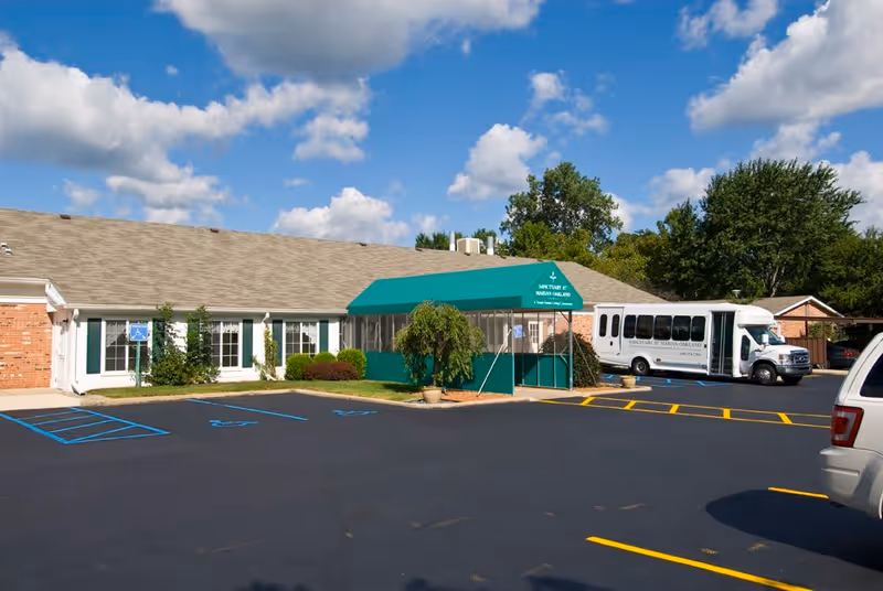Exterior view of Marian Oakland facility showing a single-story brick building with a green awning entrance. There is a parking lot with marked handicap spaces and a white shuttle bus parked near the entrance. Trees and a partly cloudy blue sky are visible in the background.