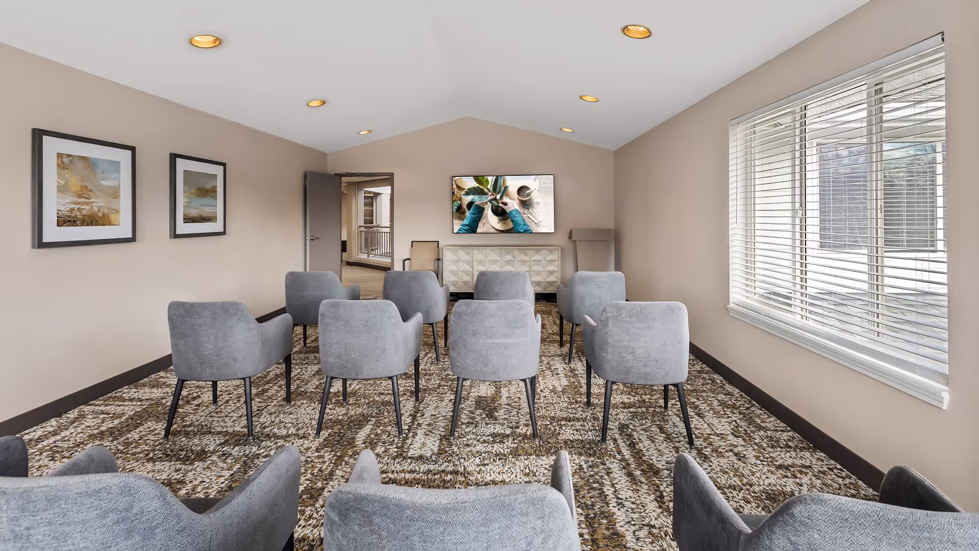 A small meeting or media room with two rows of gray upholstered chairs facing a wall-mounted flat screen TV. The room has beige walls, a patterned carpet, two framed pictures on the left wall, a window with blinds on the right, and recessed ceiling lights.