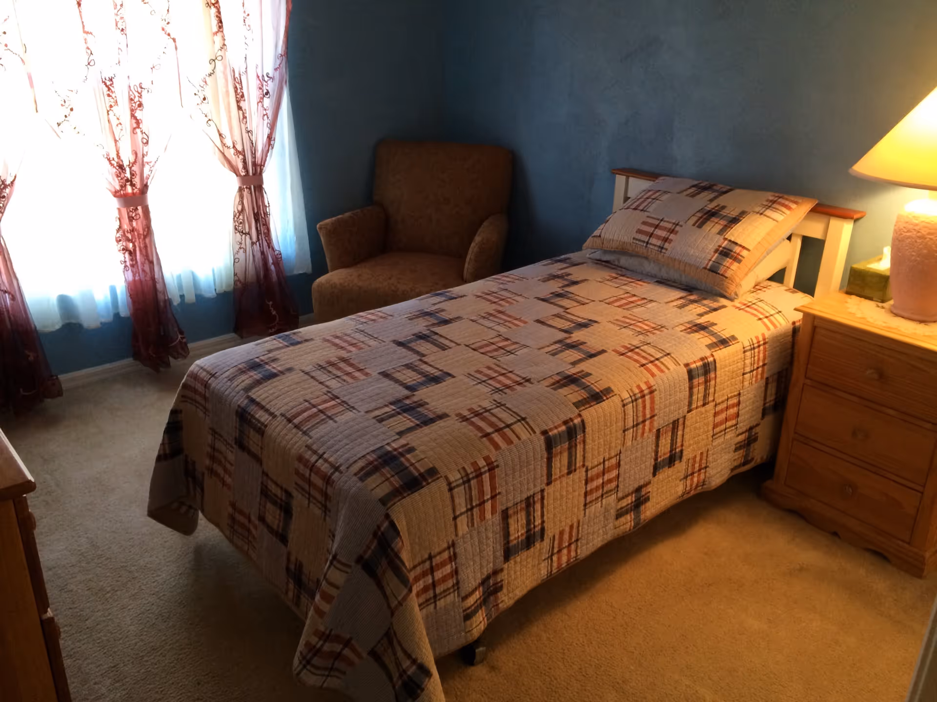 A small bedroom with a single bed covered in a patterned quilt and matching pillow. Next to the bed is a wooden nightstand with a lamp and a box of tissues. There is a cushioned armchair in the corner near a window with sheer curtains that have a floral design. The walls are painted blue and the floor is carpeted.