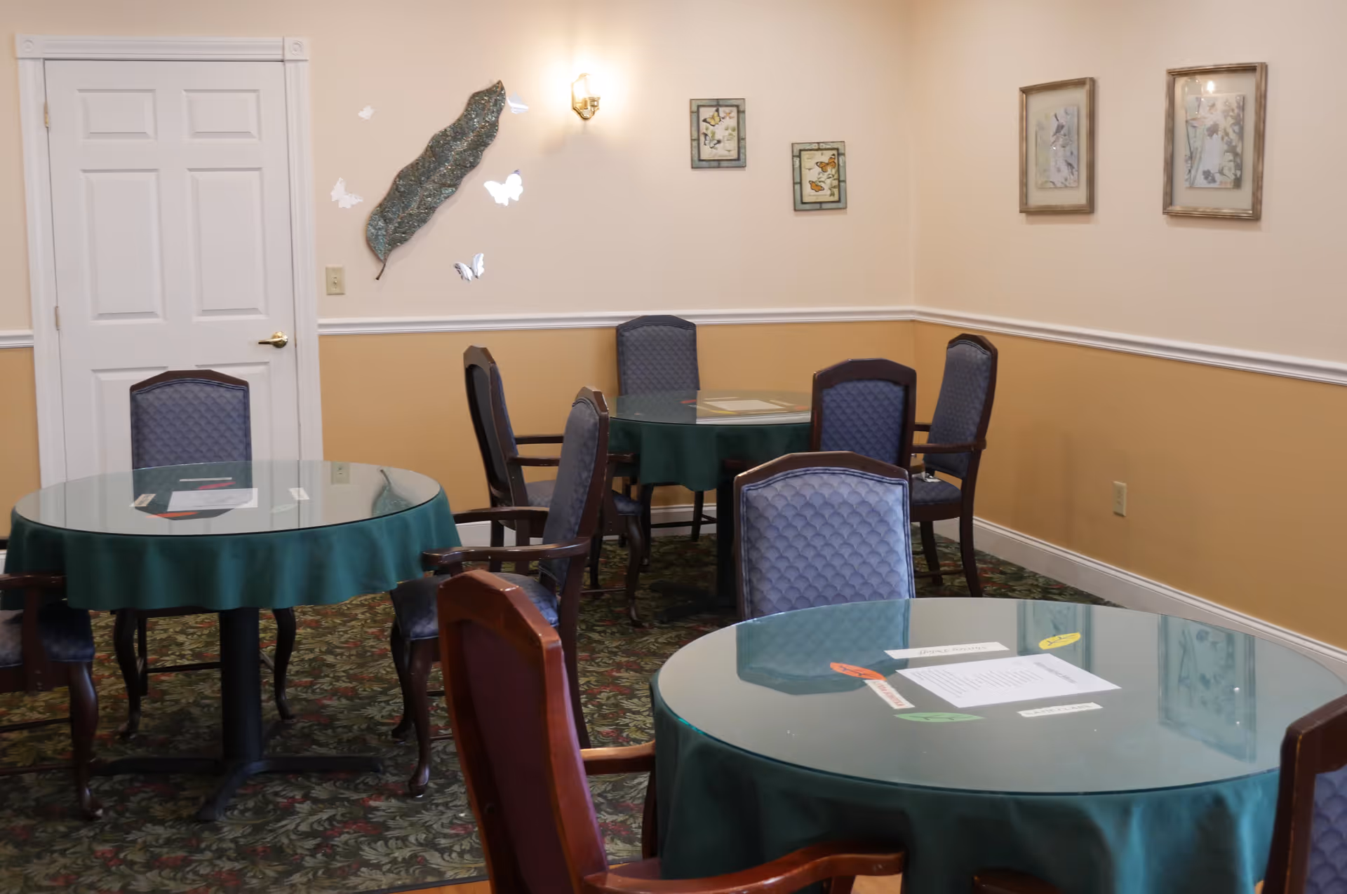 A dining room with round tables covered in green tablecloths and glass tops, surrounded by upholstered chairs. The walls are painted beige and decorated with framed pictures and wall art. The floor has a patterned carpet.