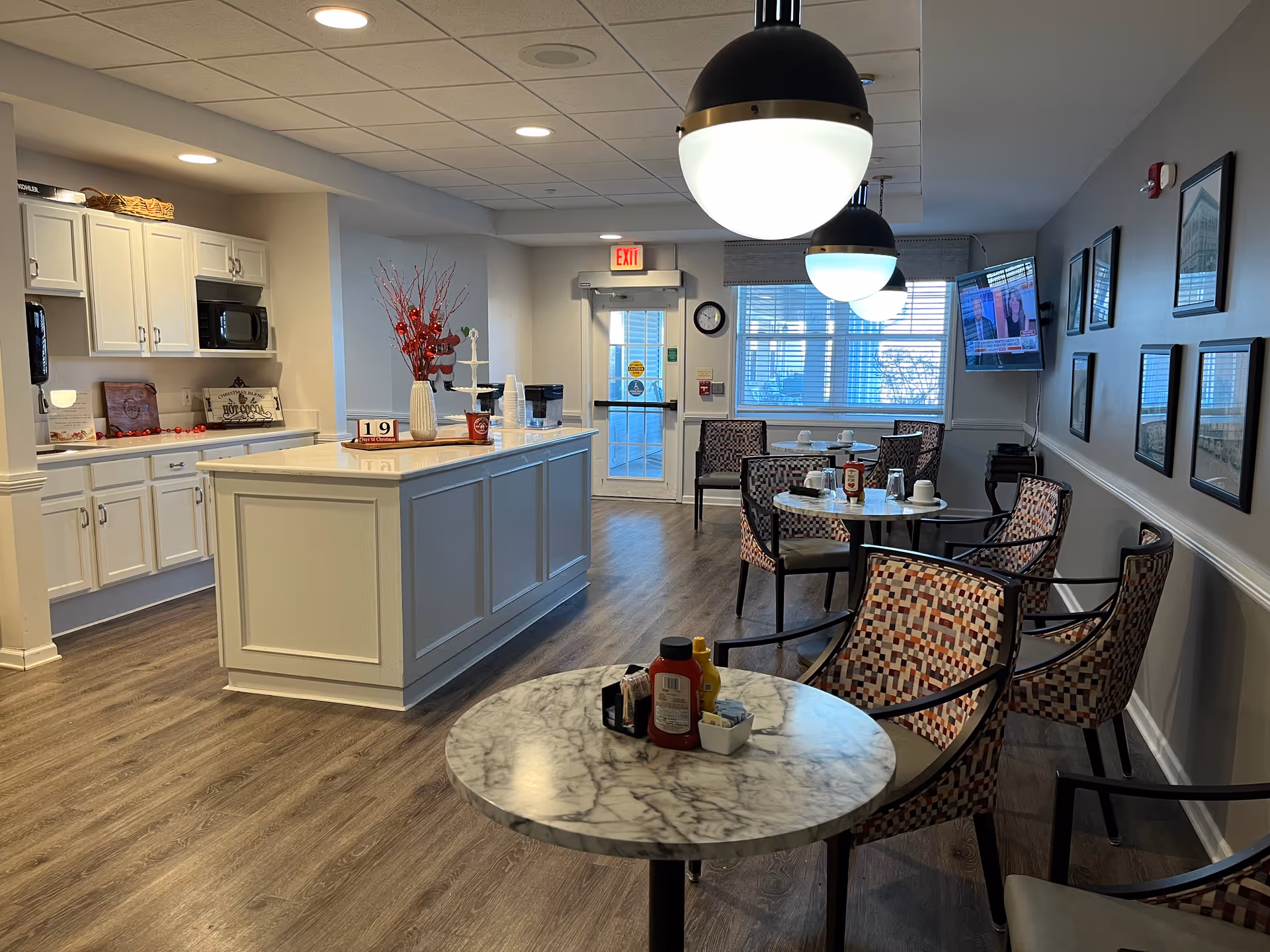 Interior view of a dining area in a senior living facility with round marble-top tables and patterned chairs. A kitchen counter with white cabinets and a microwave is visible on the left. The room is decorated with a vase of red branches and holiday decor. Pendant lights hang from the ceiling, and a TV is mounted on the right wall showing a news program. Large windows and a glass door provide natural light.