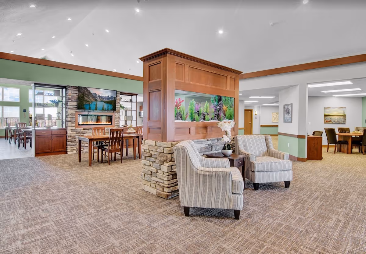 A spacious senior living facility common area featuring two striped armchairs with a small table and a potted orchid between them. Behind the chairs is a large built-in aquarium with colorful plants, set on a stone and wood base. The room has carpeted floors, green and white walls with wood trim, and recessed lighting. In the background, there is a dining area with wooden tables and chairs, a stone fireplace with a mounted TV above it, and large windows letting in natural light.