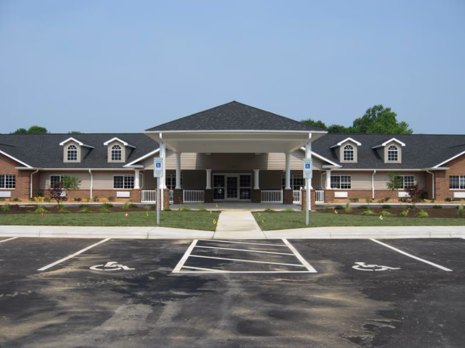 Front exterior view of Caswell House, a single-story building with a covered entrance supported by white columns, brick and beige siding, multiple windows, and a parking lot with two handicapped parking spaces in front.
