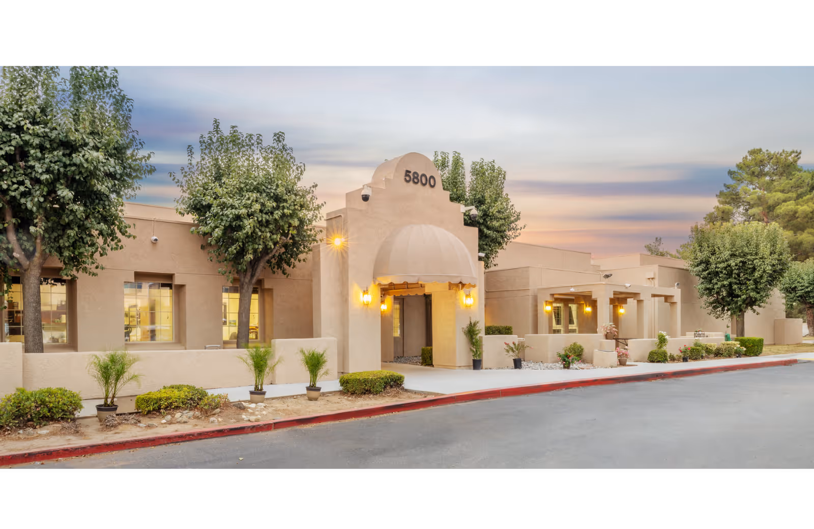 Front exterior of a single-story beige senior living facility with an arched entrance marked 5800, small trees and landscaping, and warm lights at dusk.