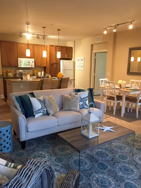 A cozy living room area with a beige sofa adorned with decorative pillows, a wooden coffee table with a white lantern and starfish decoration, and a patterned rug. Behind the sofa is an open kitchen with wooden cabinets, a white refrigerator, microwave, and three pendant lights hanging over a breakfast bar with wicker chairs. To the right, there is a dining area with a white dining table and chairs, set with plates and flowers, and a large mirror on the wall.
