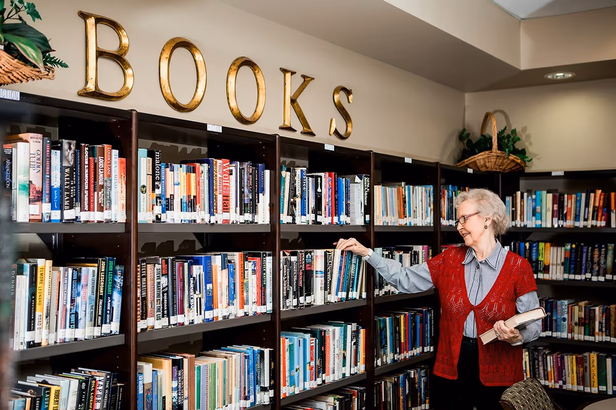 An elderly woman wearing glasses and a red knitted vest is standing in front of a bookshelf filled with books in a library area. She is holding a book in one hand and reaching for another book on the shelf. Above the bookshelf, large gold letters spell out the word 'BOOKS'. The room has a cozy and well-lit atmosphere with plants placed on top of the shelves.