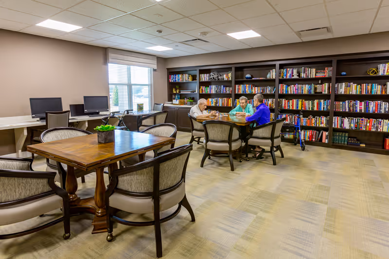 Three older adults sit at a table in a library-style common room with bookshelves, tables, and computer stations.