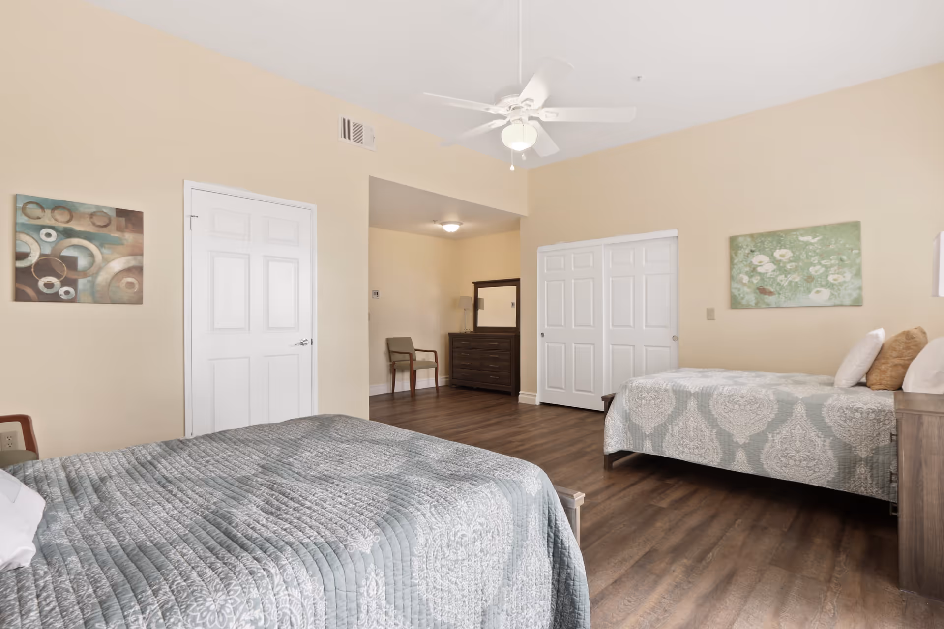 Spacious bedroom with two beds, a ceiling fan, dresser, chair, and hardwood floors in neutral decor.