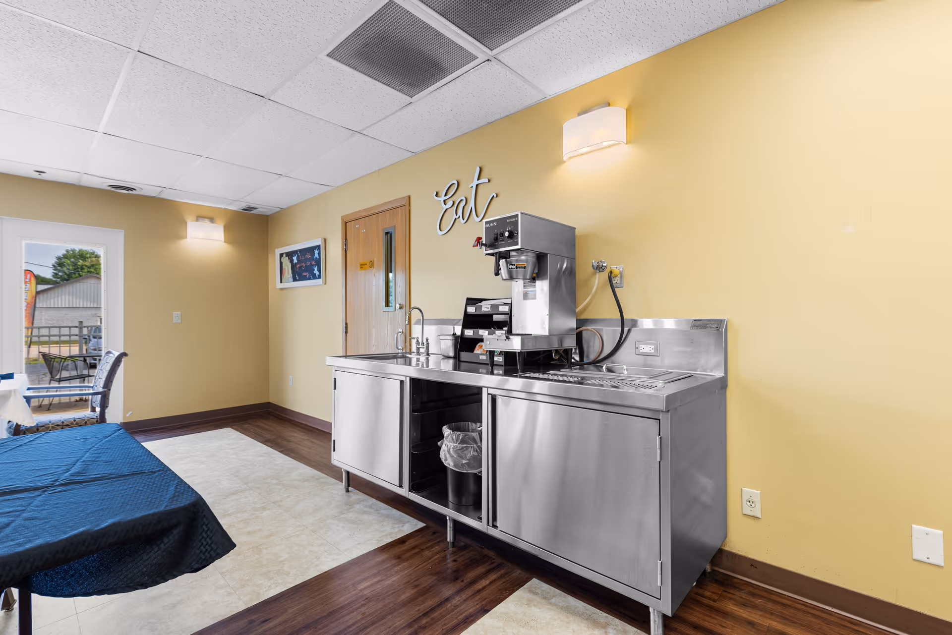 Interior view of a room with a stainless steel beverage station including a coffee machine and sink against a yellow wall. The wall has a decorative sign that says 'Eat'. There is a table covered with a dark blue tablecloth and chairs near a door leading outside to a patio area.