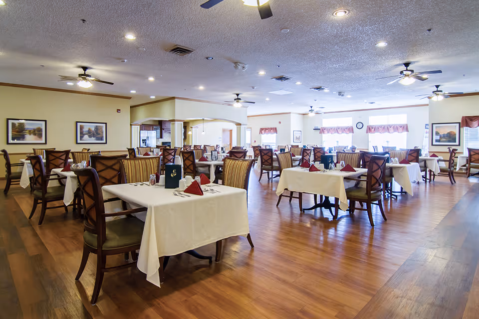 A spacious dining room with multiple tables covered in white tablecloths, each set with napkins, utensils, and condiments. The room has wooden floors, ceiling fans with lights, framed landscape paintings on the walls, and large windows with red valances allowing natural light to fill the space.