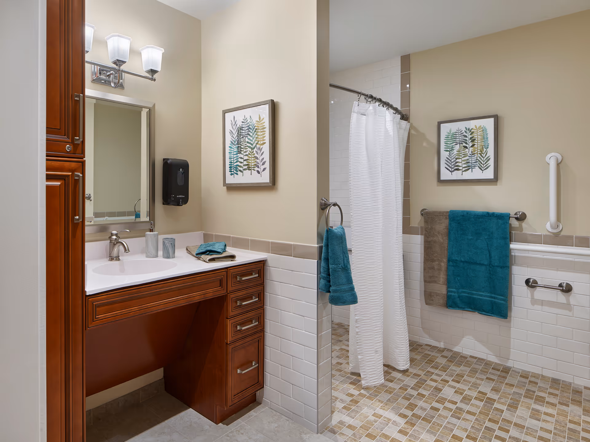 A bathroom with a wooden vanity featuring a white countertop and sink, a mirror with light fixtures above it, and a soap dispenser mounted on the wall. The bathroom has a walk-in shower with a white curtain, beige and white tiled walls, and a tiled floor in shades of beige and brown. There are two framed botanical prints on the walls and teal and brown towels hanging on towel racks. A white grab bar is mounted on the wall near the shower.