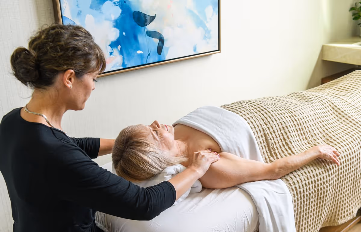 A woman receiving a shoulder massage while lying on a massage table covered with a beige textured blanket in a softly lit room with a blue abstract painting on the wall.