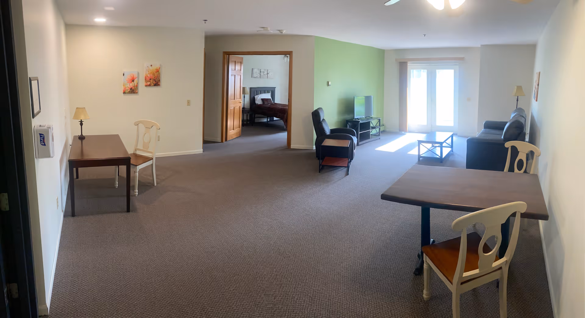 A spacious assisted living apartment with a carpeted floor, featuring a small dining table with two chairs on the right, a desk with a chair and lamp on the left, a living area with a black leather sofa, armchair, coffee table, and TV on a stand against a green accent wall. In the background, an open doorway reveals a bedroom with a bed and nightstand. French doors at the far end let in natural light.