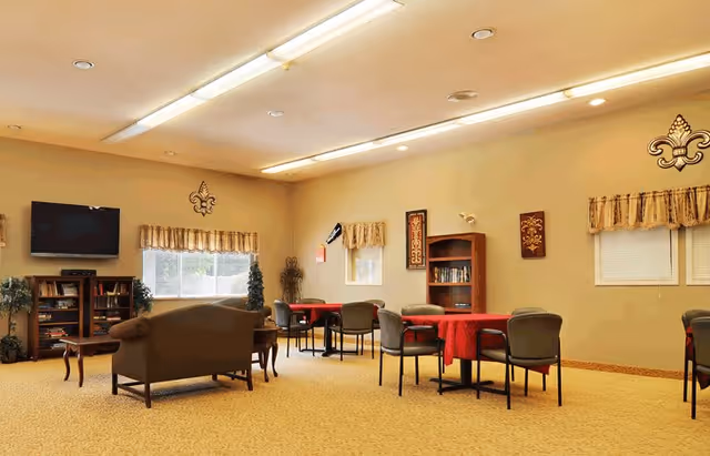 A spacious common area with beige walls and carpet, featuring several tables covered with red tablecloths and surrounded by chairs. There is a brown couch and a wooden coffee table in the foreground, a flat-screen TV mounted on the wall, bookshelves, decorative plants, and wall decorations including fleur-de-lis symbols and framed artwork. Windows have valance curtains allowing natural light to enter.