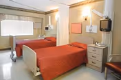 Two hospital-style beds with red bedspreads in a shared patient room with medical equipment, a nightstand, and a curtained divider.