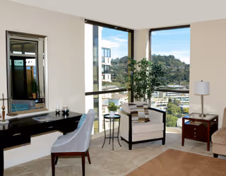 A bright living room with large corner windows offering a view of a hillside and nearby buildings. The room features a beige armchair with a striped pillow, a small round side table with a plant, a dark wooden desk with a mirror above it, a gray upholstered chair, a floor lamp, and a beige carpet.