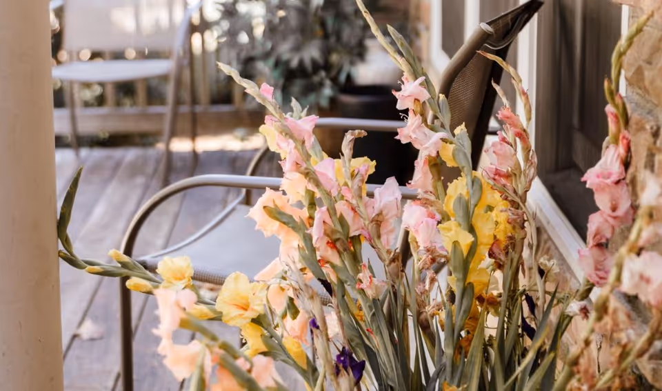 Close-up view of a bunch of colorful gladiolus flowers in shades of pink, yellow, and purple placed near a porch with metal chairs and a wooden floor. A stone wall and window frame are visible in the background.