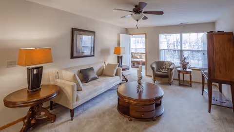 A cozy living room with a beige sofa adorned with cushions, a round wooden coffee table with decorative candles, a side table with a lamp, an armchair near a window, and a ceiling fan above. The room is well-lit with natural light coming through the windows.