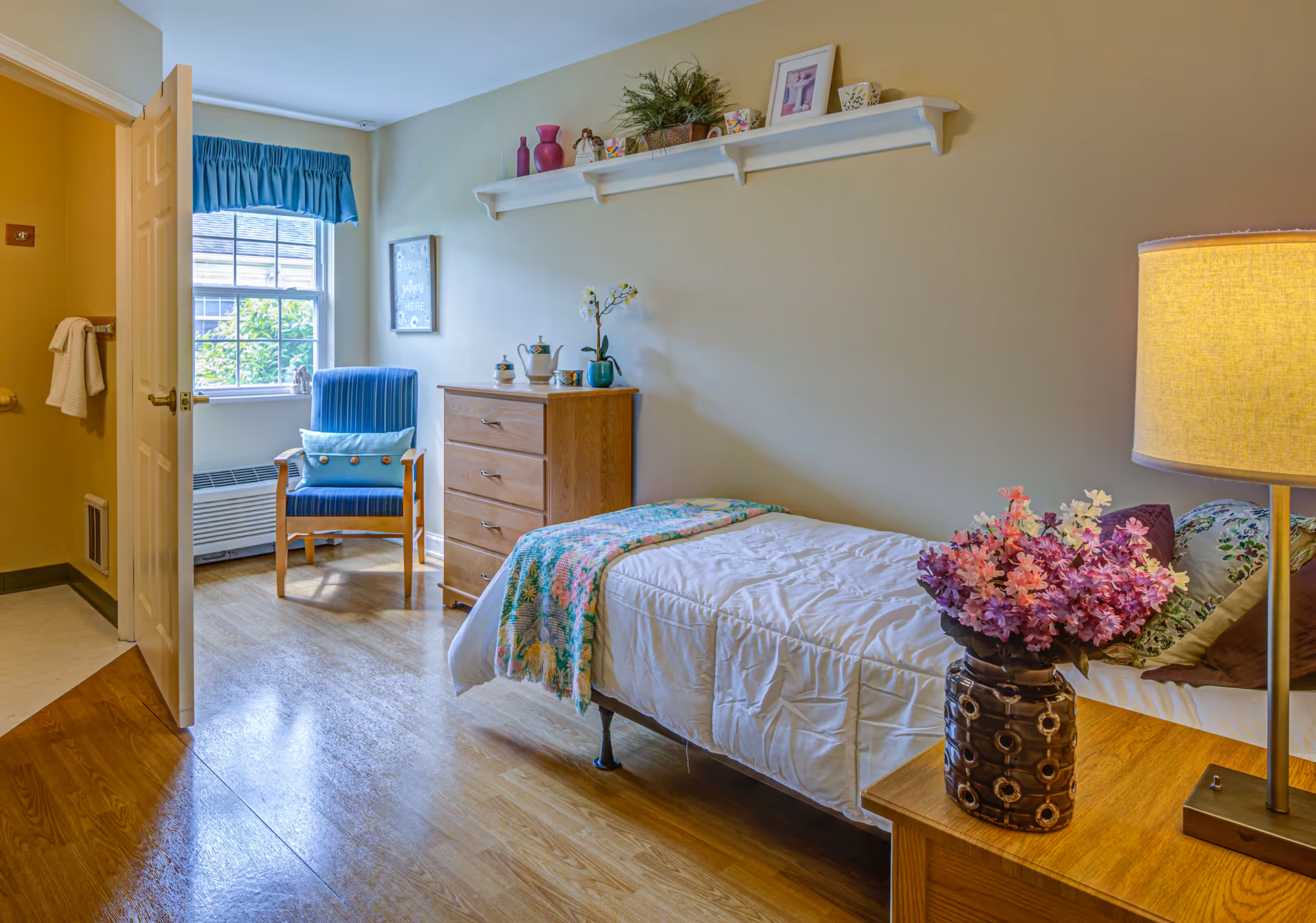 A bright, neatly arranged single-occupancy bedroom with a bed, nightstand and lamp, floral arrangement, dresser and an armchair by a window.