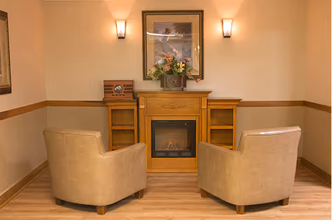 A cozy interior room with two beige armchairs facing a wooden electric fireplace. Above the fireplace is a framed picture and a floral arrangement. Two wall sconces with warm lighting flank the picture. The room has light brown walls with a darker brown chair rail and wood flooring.