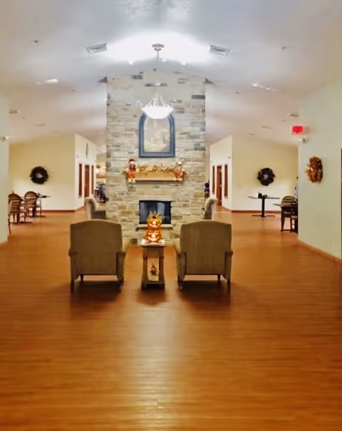 A spacious senior living common area with wooden flooring, two armchairs facing a stone fireplace decorated with autumn-themed ornaments, and a chandelier hanging from the ceiling. There are tables and chairs along the walls, wreaths hanging on the walls, and soft lighting creating a warm atmosphere.