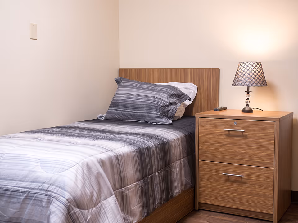 A single bed with a wooden headboard and gray striped bedding next to a wooden nightstand with two drawers. On the nightstand, there is a lamp with a patterned lampshade and a remote control. The room has plain light-colored walls.