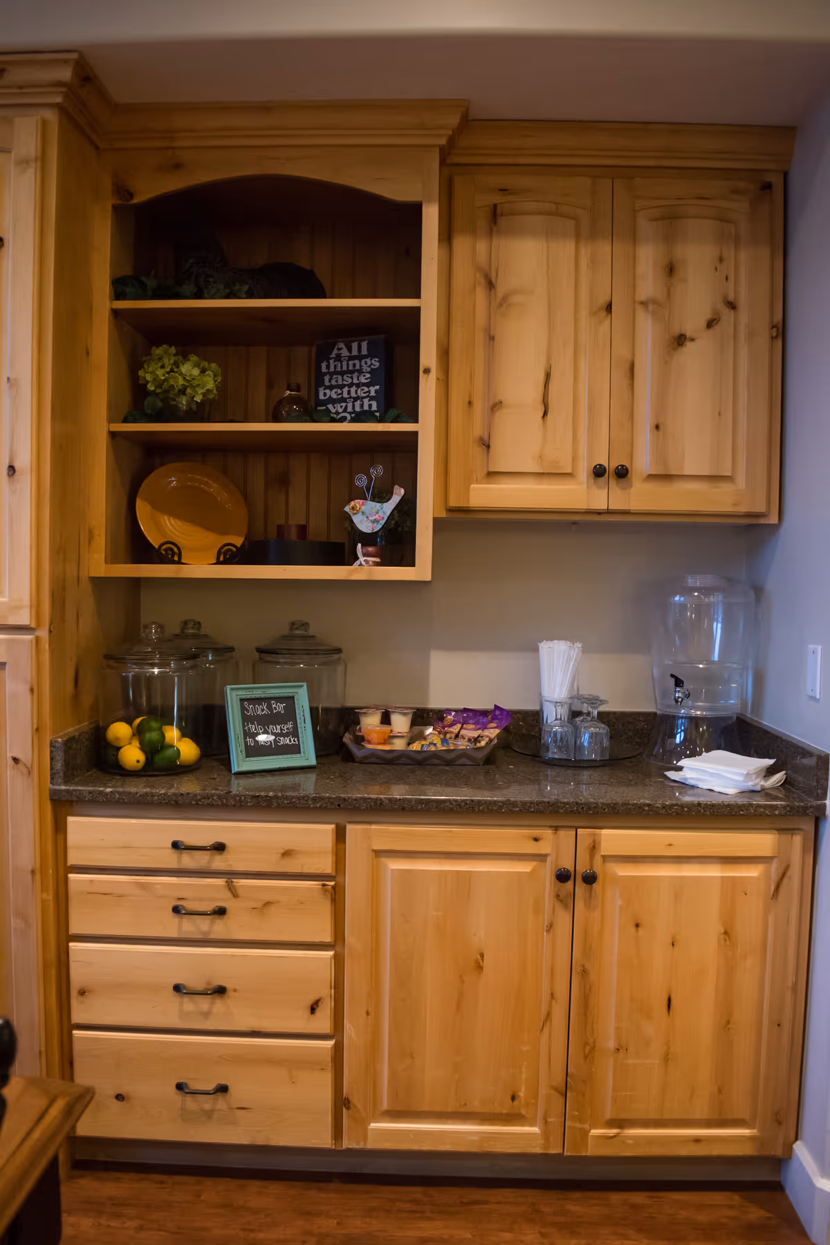 Wooden kitchenette with open shelves and cabinets above a countertop holding glass jars of fruit, a small snack display, utensils, and a water dispenser.