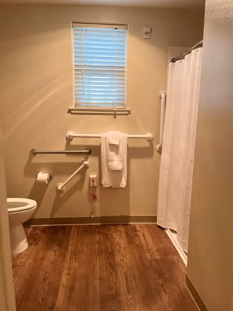A bathroom with wooden flooring, a toilet on the left, grab bars on the walls, a towel hanging on a horizontal grab bar, a window with blinds above the towel, and a shower with a white curtain on the right.