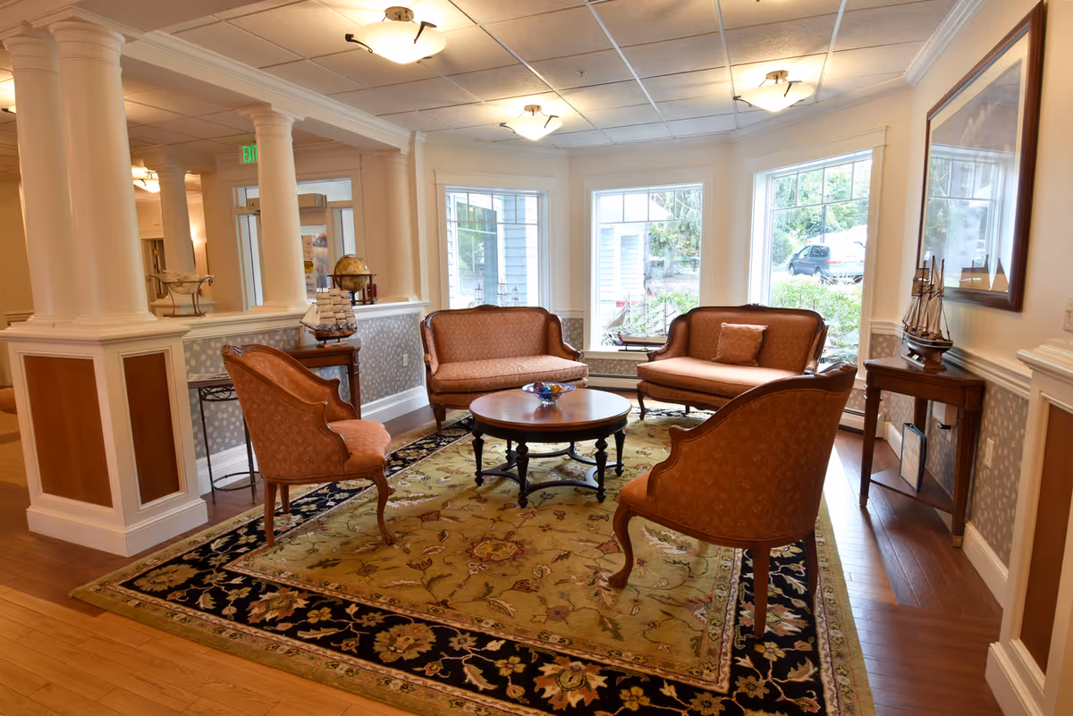 Sunlit seating area with upholstered chairs and sofas arranged around a round coffee table on a patterned rug.