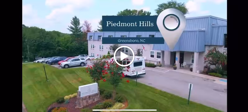 Exterior view of Piedmont Hills Center for Nursing and Rehabilitation in Greensboro, NC, showing a multi-story building with a blue roof, a parking lot with several cars, a white van, green lawn, and landscaped bushes and trees.