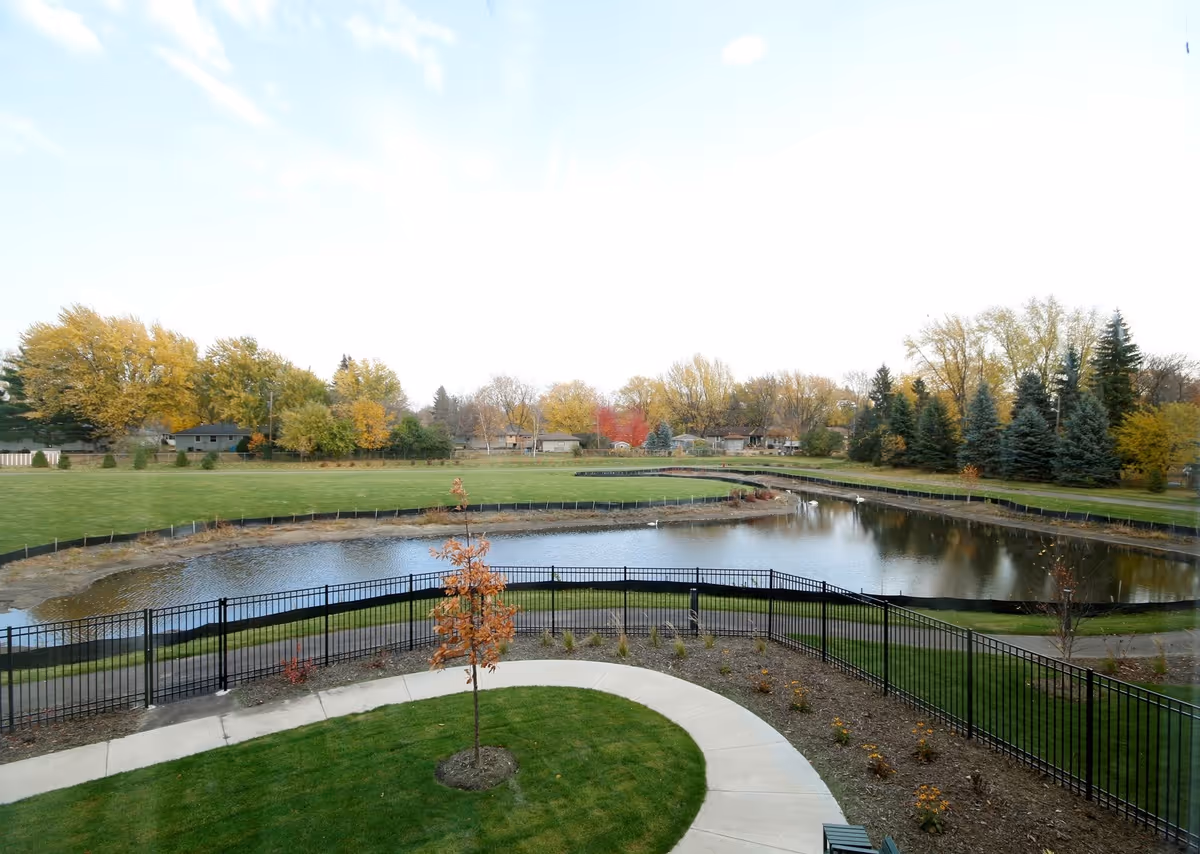 View of a landscaped outdoor area with a small pond surrounded by a black metal fence, green grass, a curved concrete walkway, and trees with autumn foliage in the background under a partly cloudy sky.