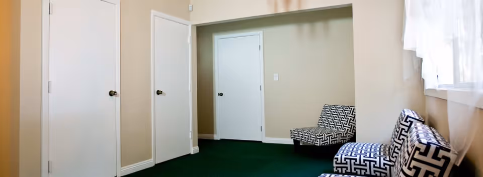 A small waiting or sitting area with three black and white patterned chairs along the right wall, beige walls, green carpet, three white doors on the left and center walls, and a window with sheer white curtains on the right.