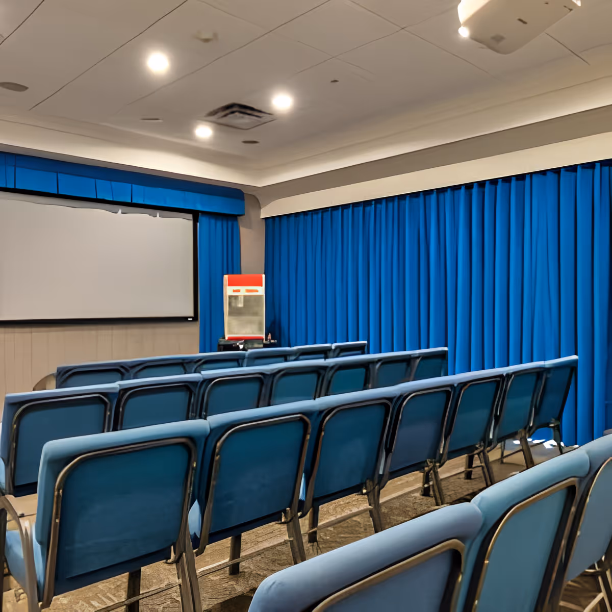 A small theater room with rows of blue cushioned chairs facing a large white projection screen. Blue curtains cover the walls, and a popcorn machine is visible near the screen.