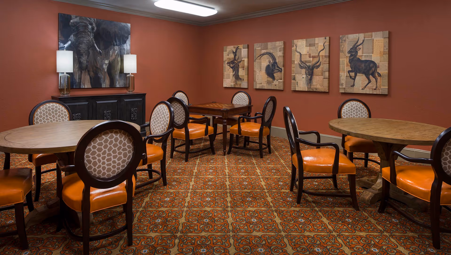 A cozy dining area with round wooden tables and cushioned chairs featuring orange seats and patterned backs. The walls are painted a warm reddish-brown color and decorated with four framed animal paintings and a large elephant painting above a dark wooden sideboard with two lamps.