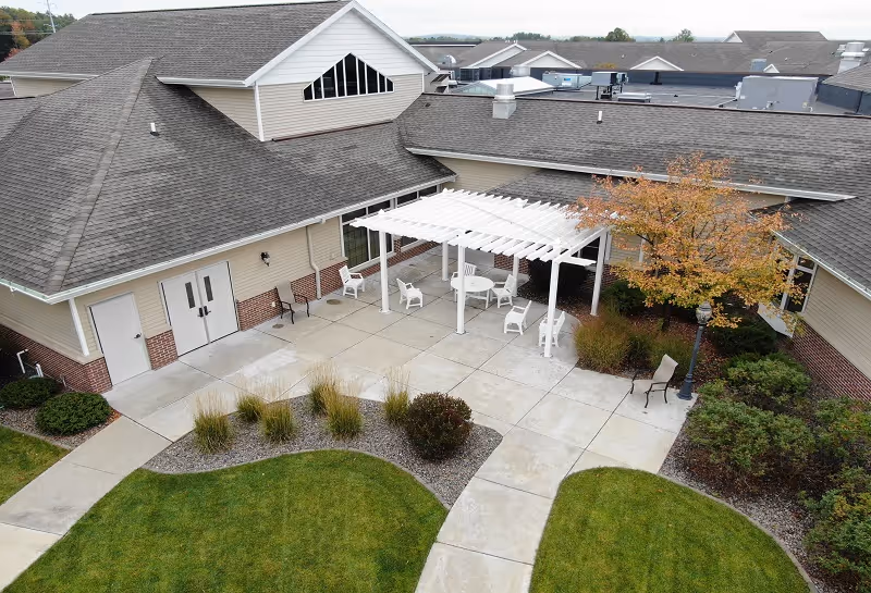 Outdoor courtyard area of a senior living facility with a white pergola, several white chairs and a table underneath. The courtyard is surrounded by beige buildings with gray roofs, landscaped bushes, a tree with autumn-colored leaves, and concrete walkways.