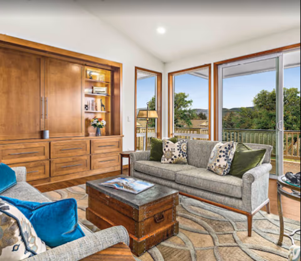 A bright living room with large windows and sliding glass door showing an outdoor view with trees and hills. The room features a gray sofa with patterned and green pillows, a wooden chest used as a coffee table with a magazine on top, a gray armchair with blue pillows, a wooden built-in cabinet with shelves holding decorative items, and a patterned area rug on a wooden floor.