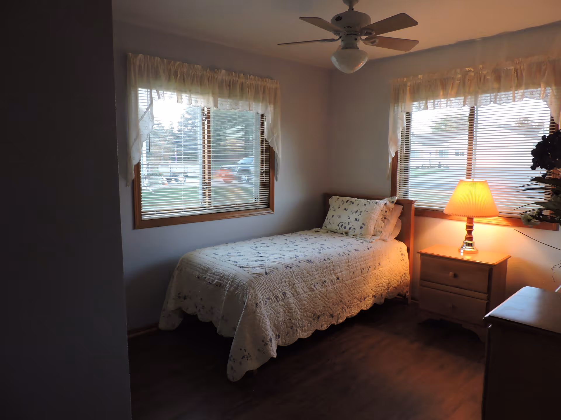 A small bedroom with a single bed covered in a white quilt with floral patterns. There are two windows with blinds and lace curtains, a wooden nightstand with a lit lamp, a ceiling fan, and wooden flooring.
