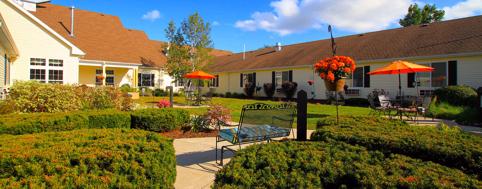 Sunny landscaped courtyard with benches, patio tables, orange umbrellas and potted flowers in front of a single-story building.