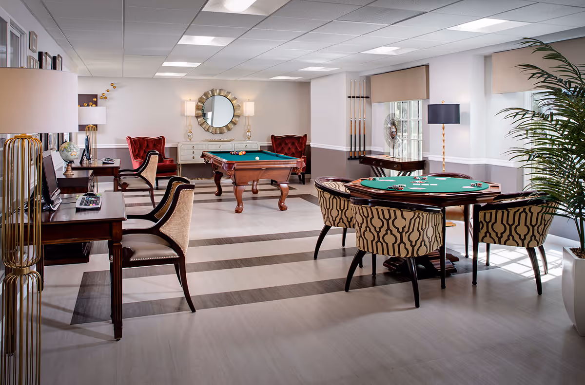 A bright and spacious game room with a pool table in the center, a poker table with four patterned chairs, two red armchairs, and two desks with chairs and desktop computers. The room has large windows with beige blinds, a decorative round mirror on the wall, several lamps, and a tall potted plant in the corner.