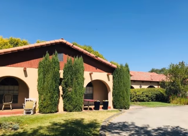Senior living facility exterior with arches, stucco walls, red-tile roof, and tall evergreen trees.